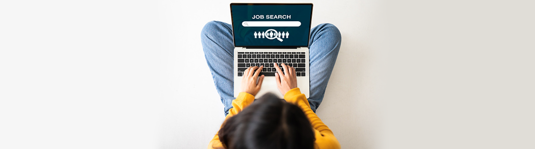 An overhead view of a woman sitting with a laptop. The laptop has a job search website open.